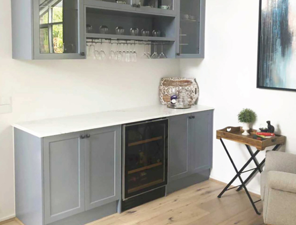 Bar area with gray cabinets, wine glasses, and a small fridge. Art on the wall and a side table with decor beside a sofa. Bar area with gray cabinets, wine glasses, and a small fridge. Art on the wall and a side table with decor beside a sofa.