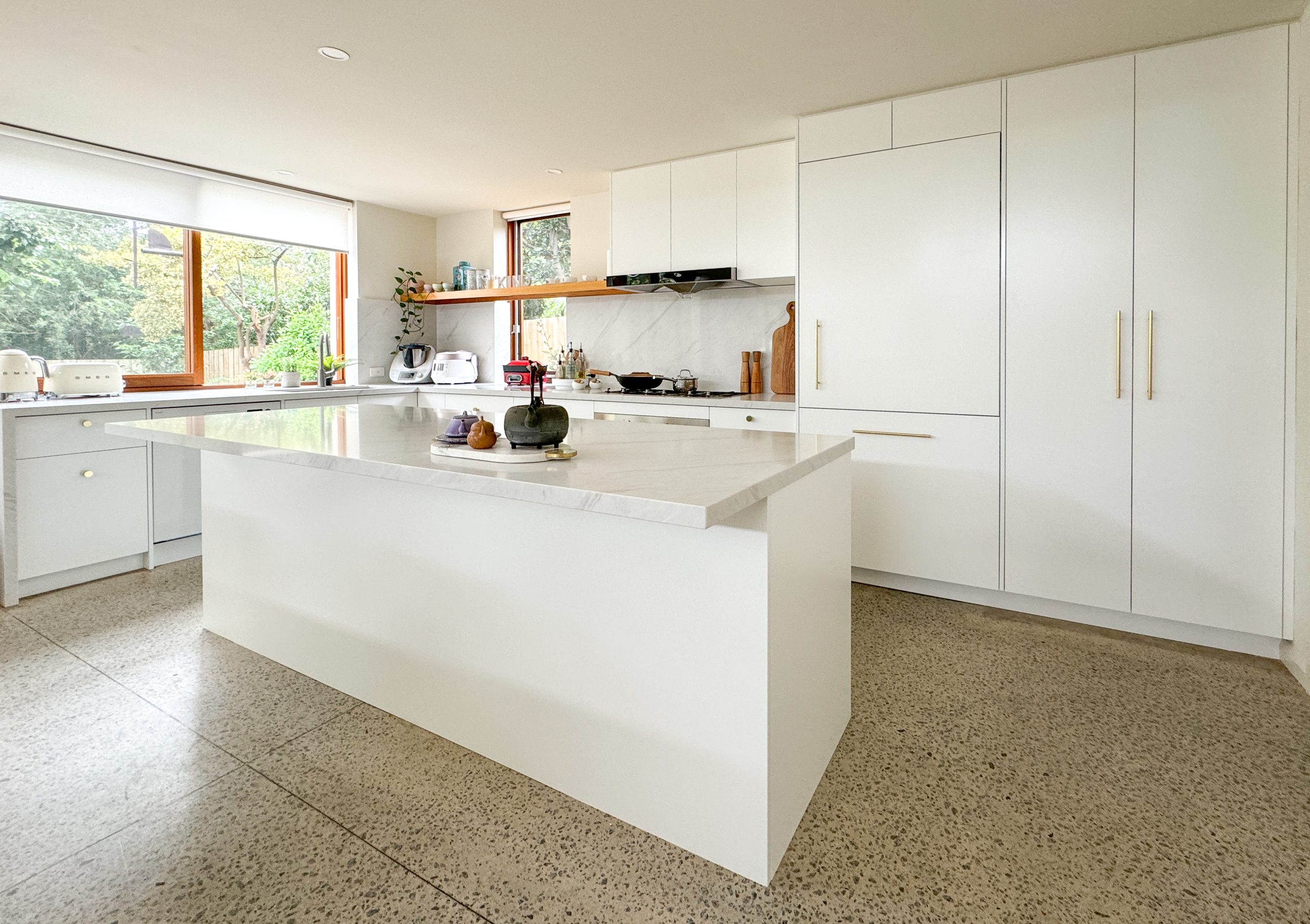 Modern kitchen with a large white island, marble countertops, and greenery visible through the windows.