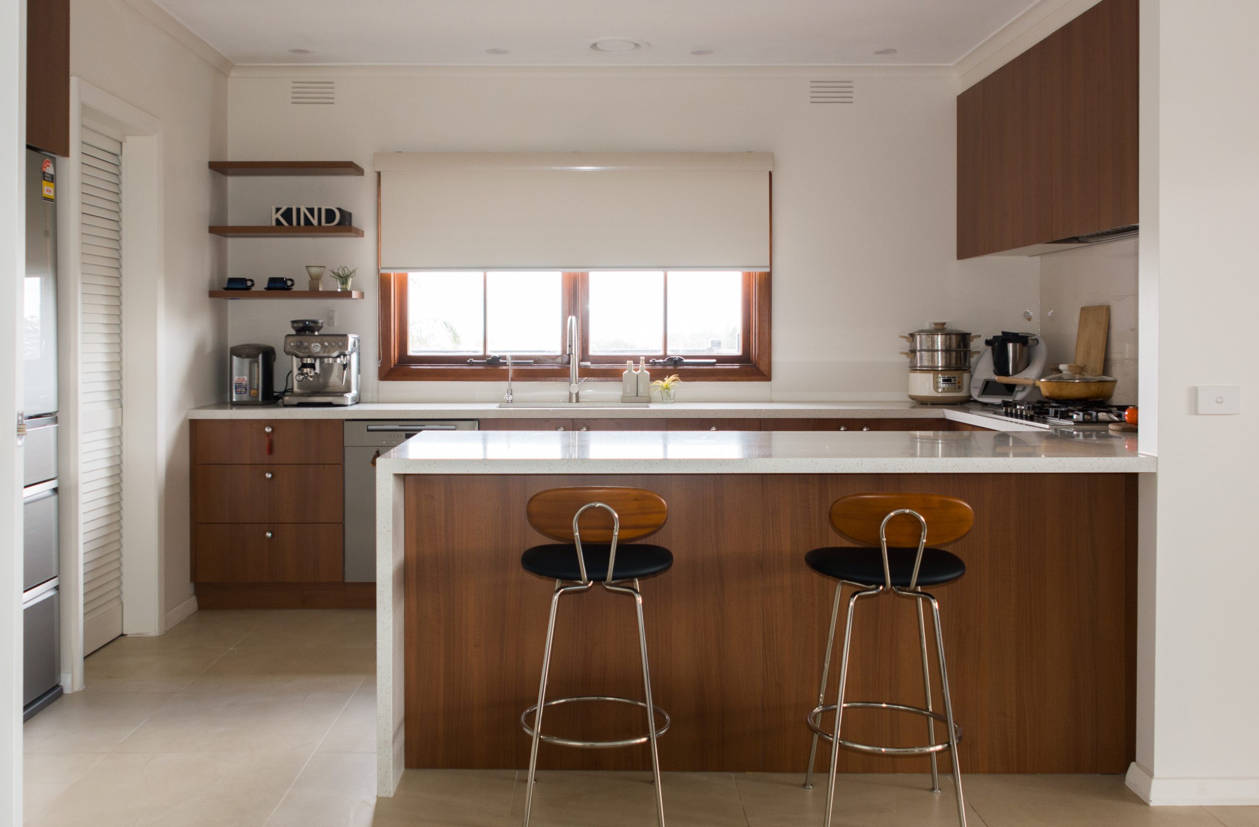 Modern kitchen with wooden cabinets, two bar stools, coffee maker, mixer, and sunlight from a window above the sink.