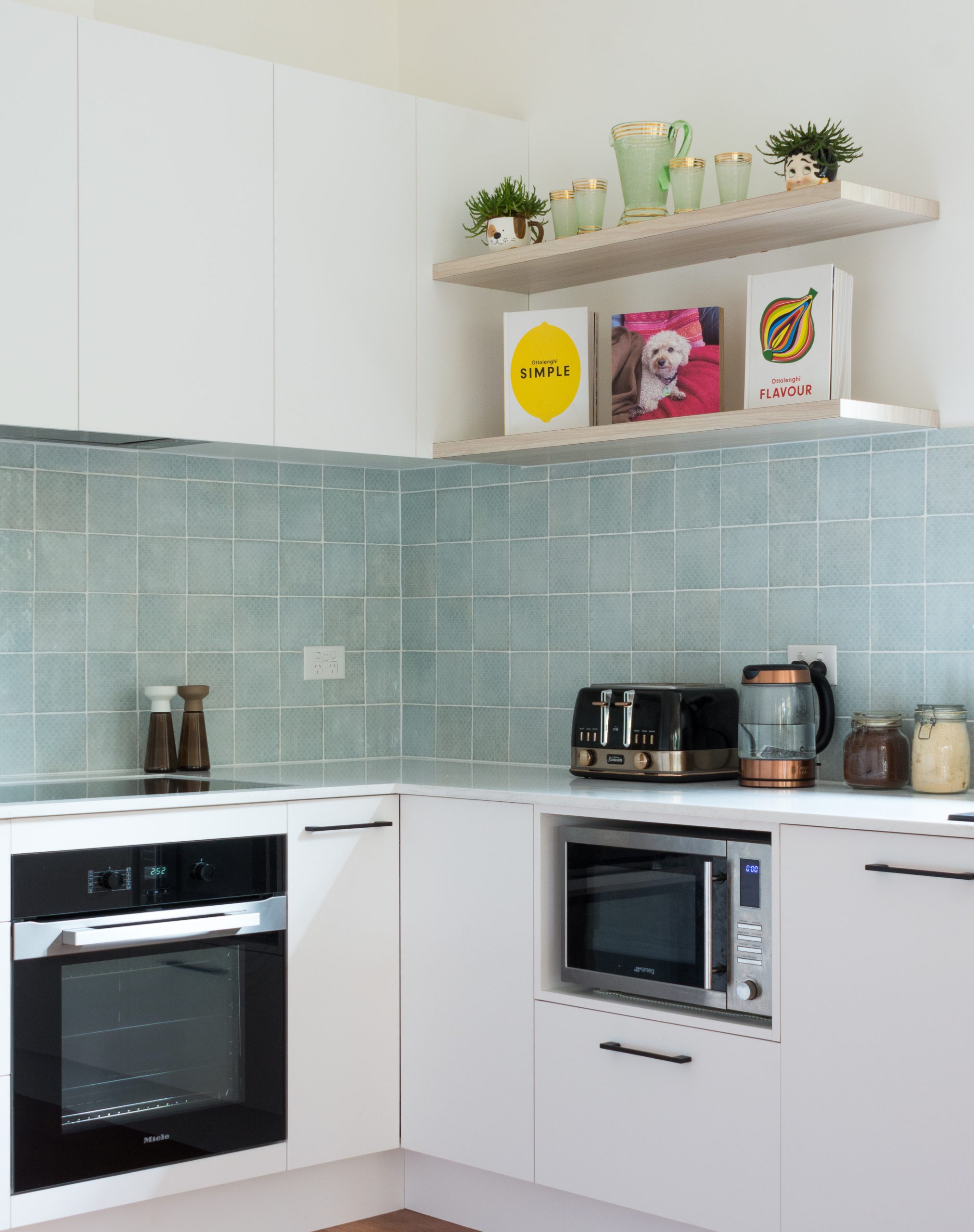 Modern kitchen with white cabinets, black appliances, teal tile backsplash, and open shelves holding plants and books. Modern kitchen with white cabinets, black appliances, teal tile backsplash, and open shelves holding plants and books.
