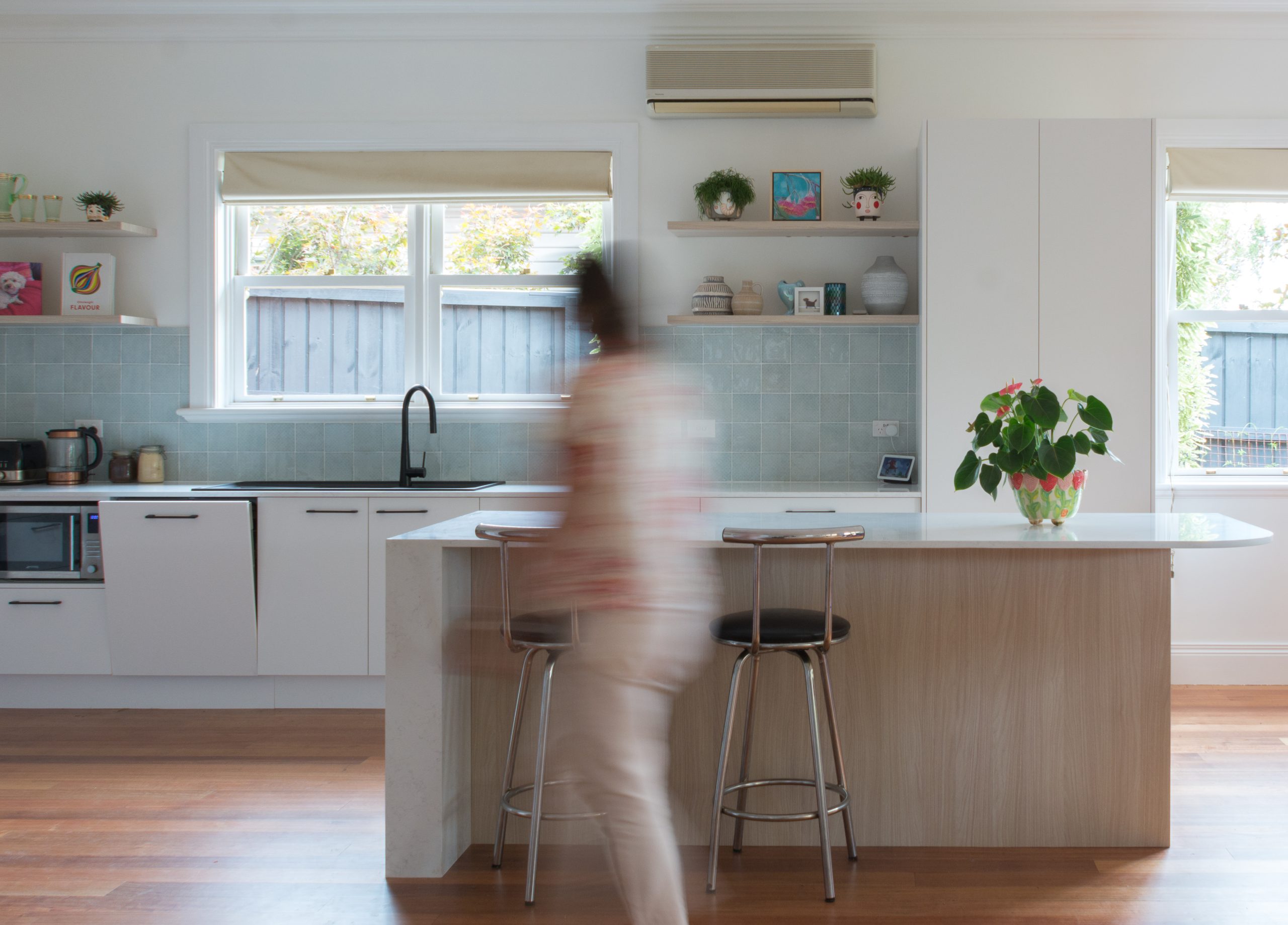 A person walking in a modern kitchen with white cabinets, bar stools, and plants on the counter. A person walking in a modern kitchen with white cabinets, bar stools, and plants on the counter.