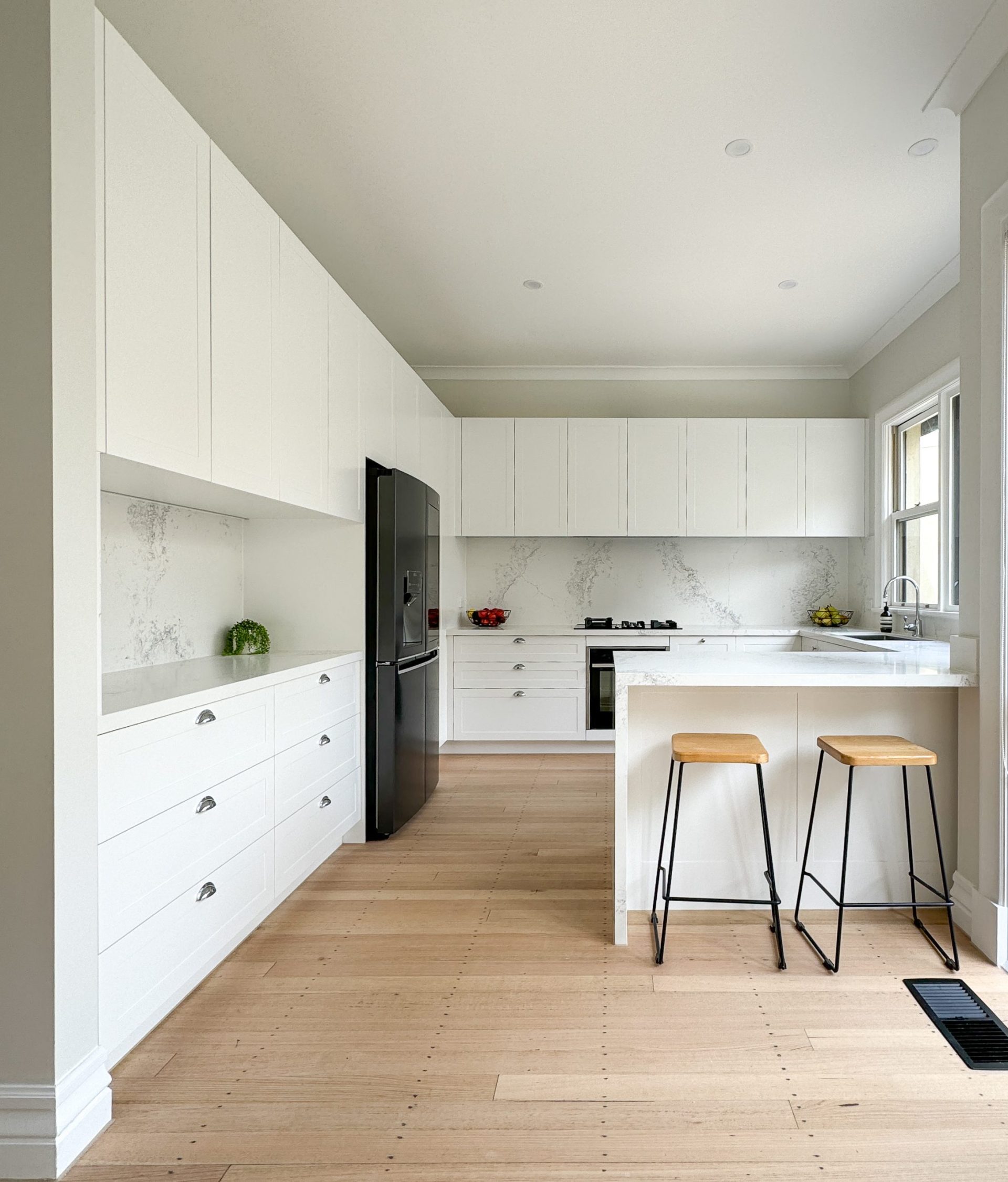 Modern kitchen with white cabinets, black fridge, light wood floors, and two wooden stools at a marble island. Modern kitchen with white cabinets, black fridge, light wood floors, and two wooden stools at a marble island.