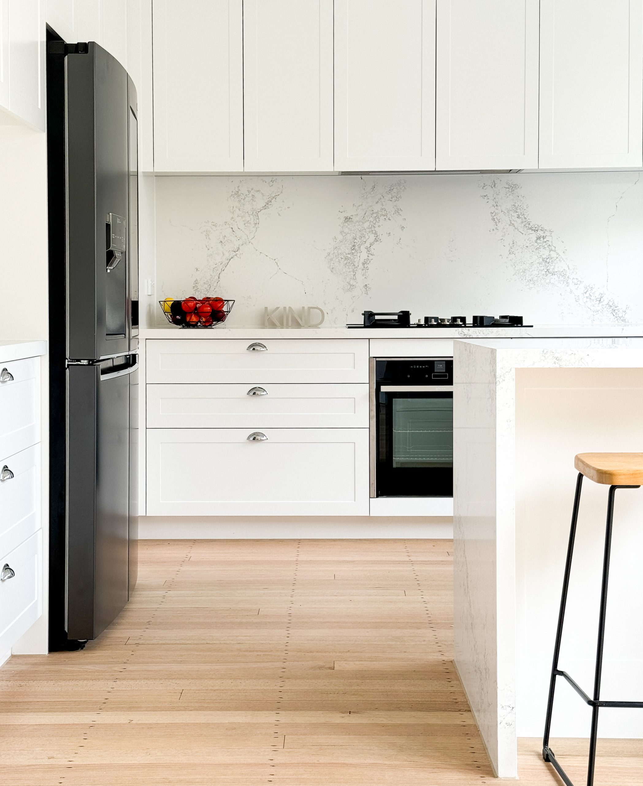 Modern white kitchen with a black fridge, marble backsplash, fruit bowl on counter, and a bar stool. Modern white kitchen with a black fridge, marble backsplash, fruit bowl on counter, and a bar stool.