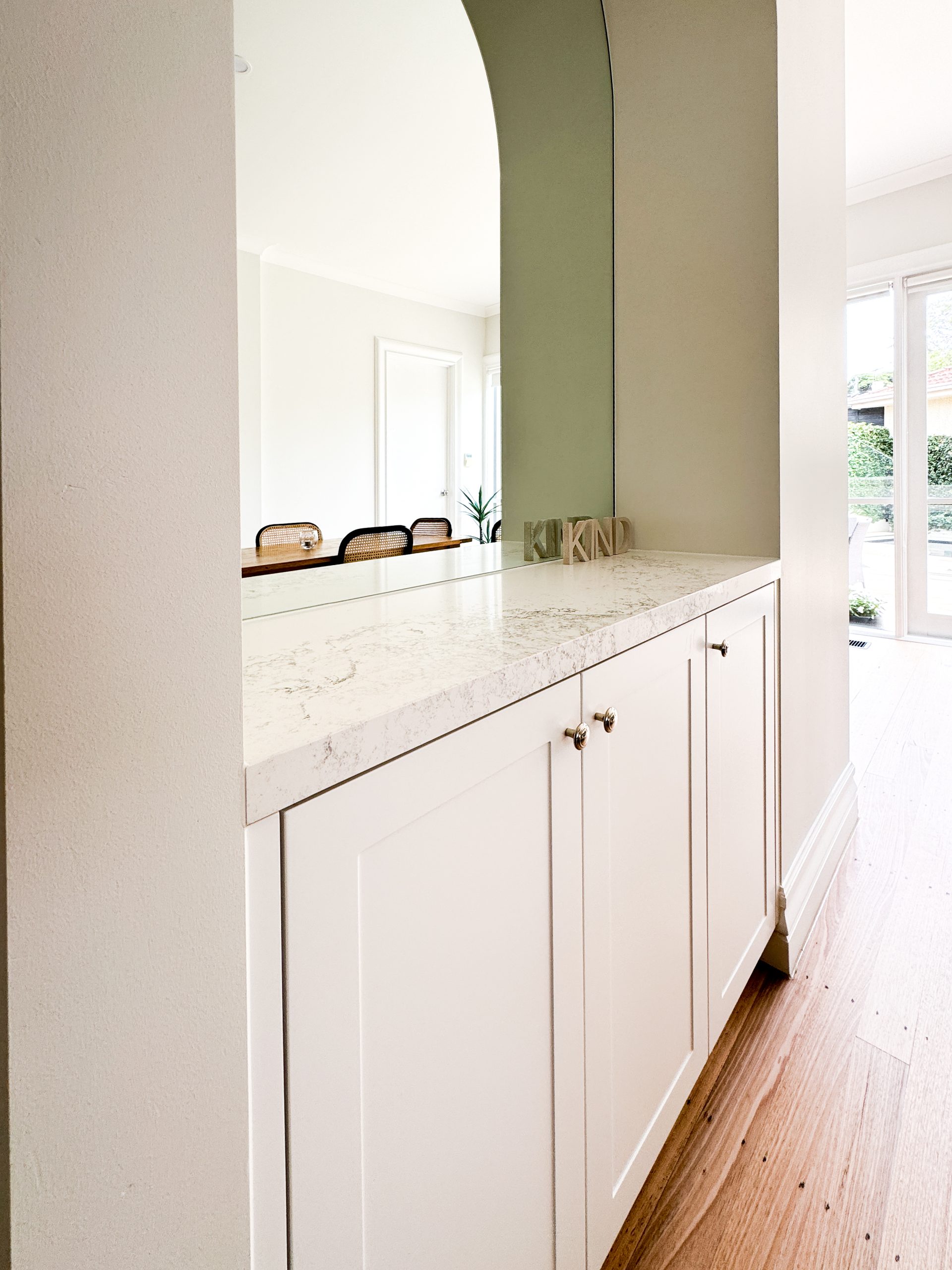White cabinet with marble countertop under an archway, leading to a bright room with large windows and chairs. White cabinet with marble countertop under an archway, leading to a bright room with large windows and chairs.