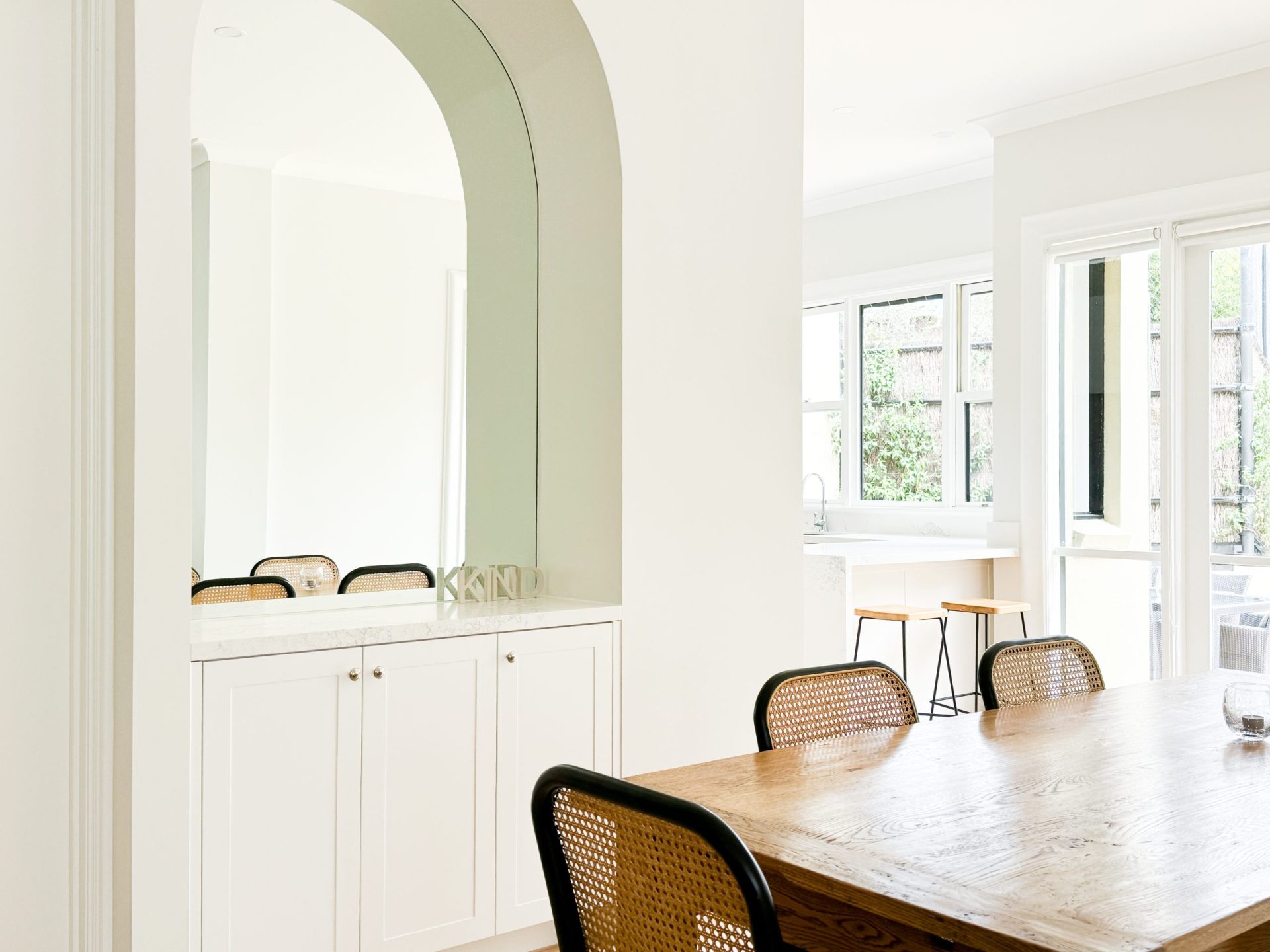 Modern dining room with wooden table, black chairs, and archway mirror. Bright natural light from large windows. Modern dining room with wooden table, black chairs, and archway mirror. Bright natural light from large windows.