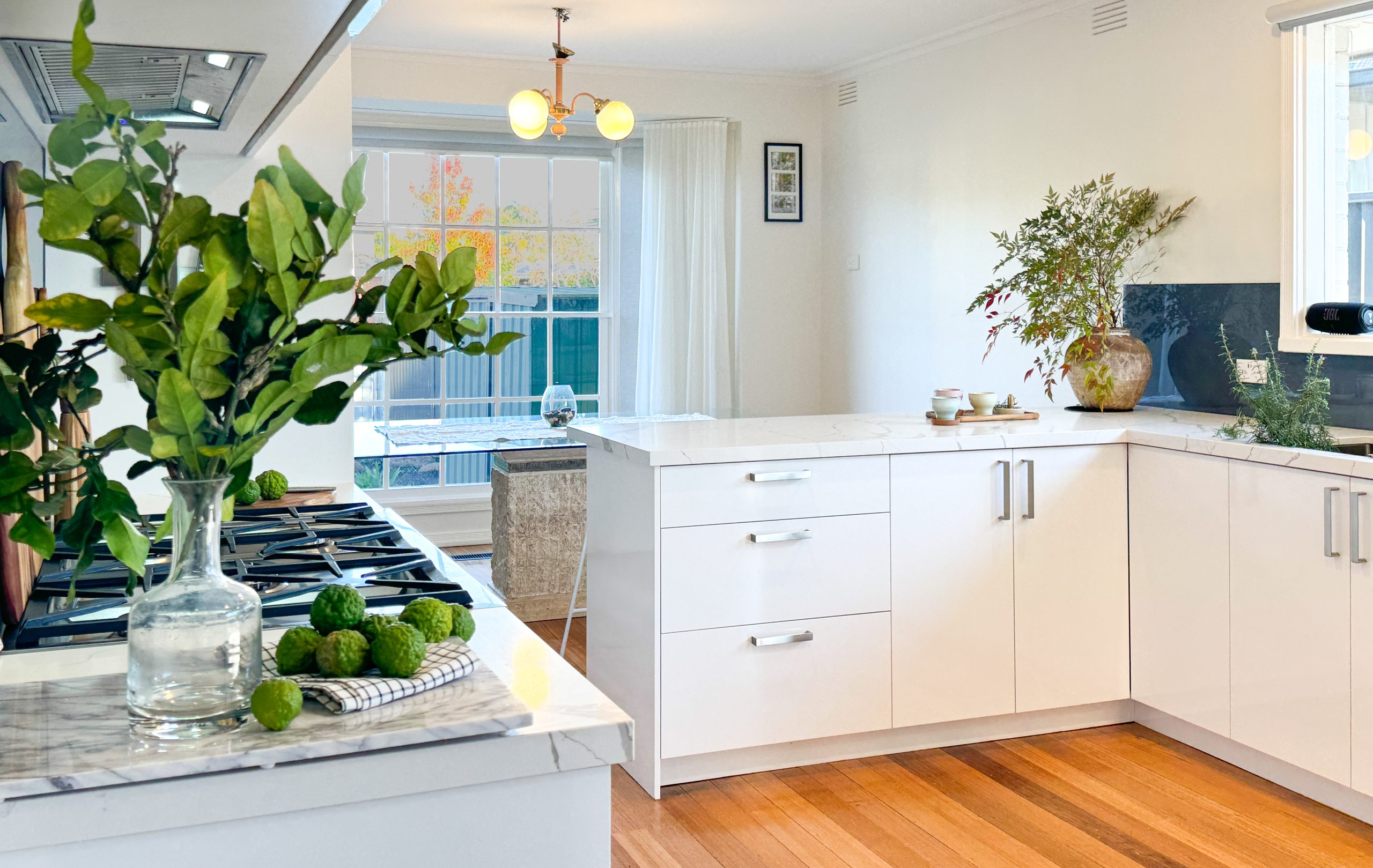 Bright kitchen with white cabinets, marble countertops, green plants, and wooden flooring, next to a sunlit dining area.