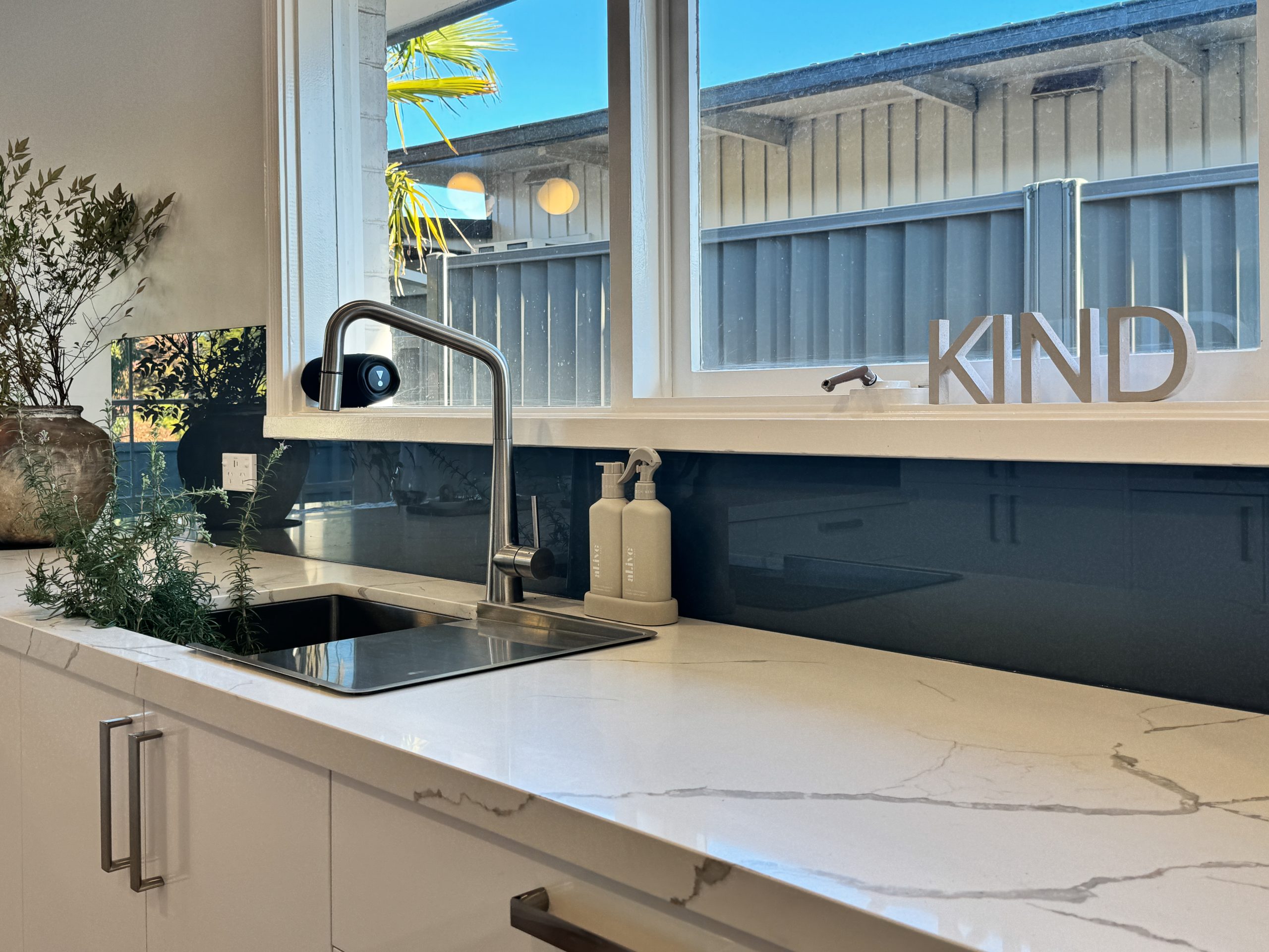 Modern kitchen with marble counter, sink, soap dispensers, plants, and “KIND” letters by a large window. Modern kitchen with marble counter, sink, soap dispensers, plants, and "KIND" letters by a large window.