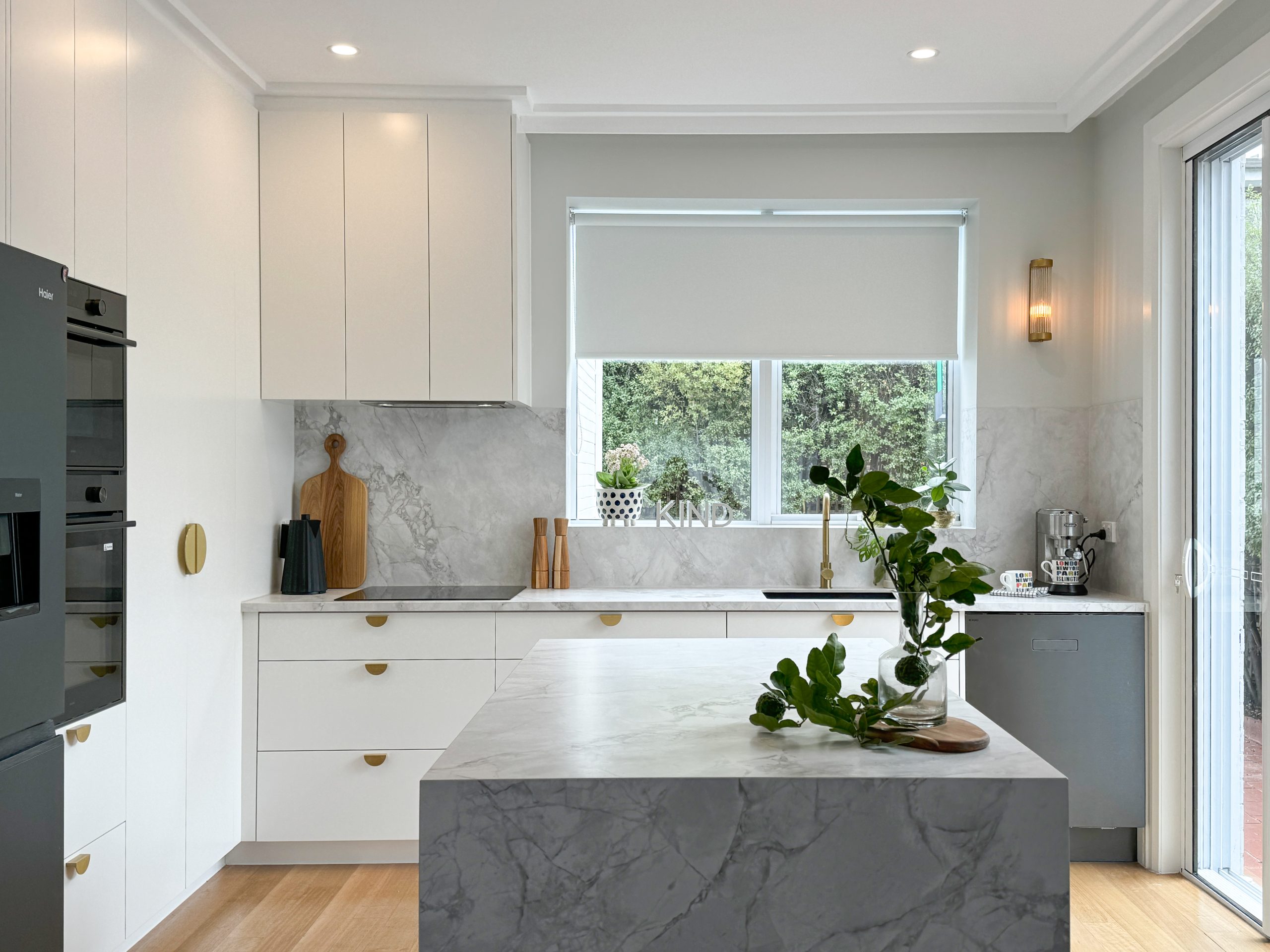 Modern kitchen with marble island, white cabinets, and green plants. Large window overlooks greenery. Modern kitchen with marble island, white cabinets, and green plants. Large window overlooks greenery.