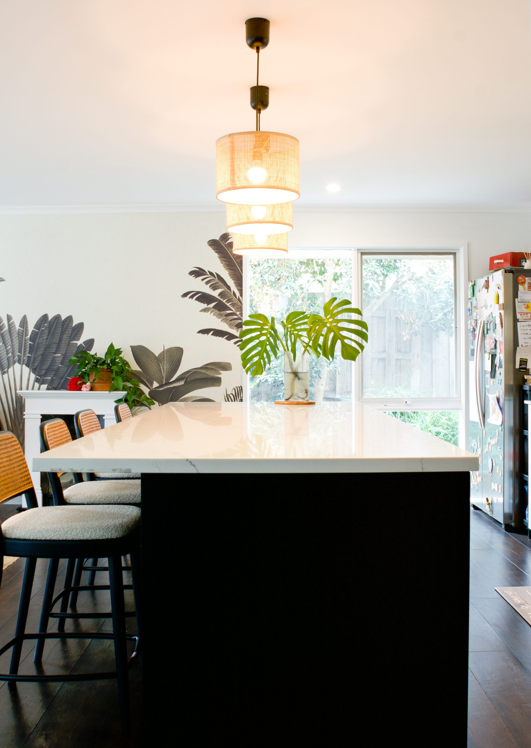 Modern kitchen with a white island countertop, stools, leafy plants, large window, and pendant lighting. Modern kitchen with a white island countertop, stools, leafy plants, large window, and pendant lighting.