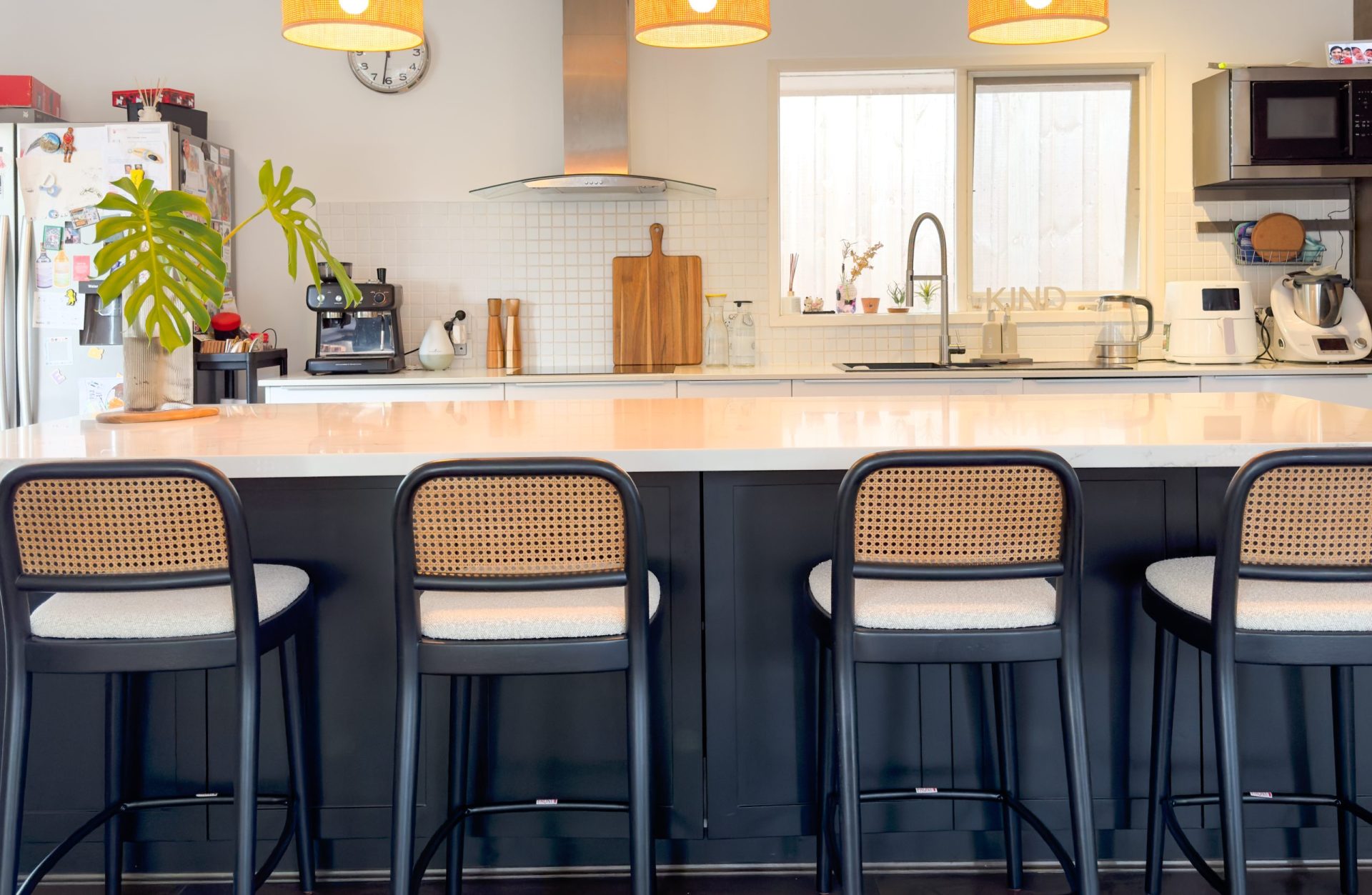 Modern kitchen with a white island, dark stools, hanging lamps, and appliances, featuring a plant and wooden accents. Modern kitchen with a white island, dark stools, hanging lamps, and appliances, featuring a plant and wooden accents.