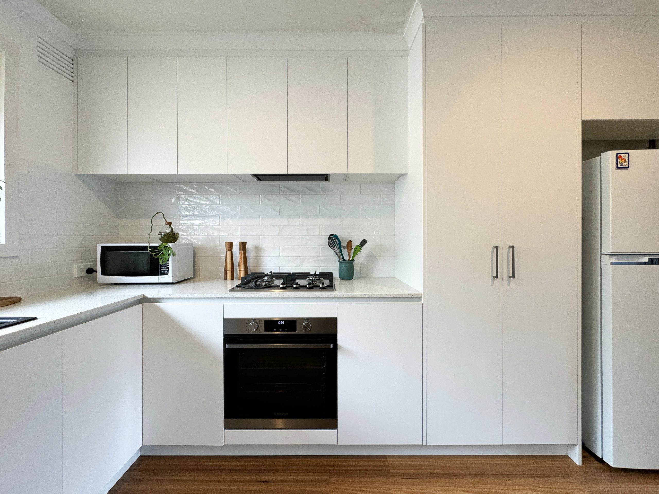 Modern white kitchen with oven, microwave, stove, knife block, and refrigerator on wooden flooring.