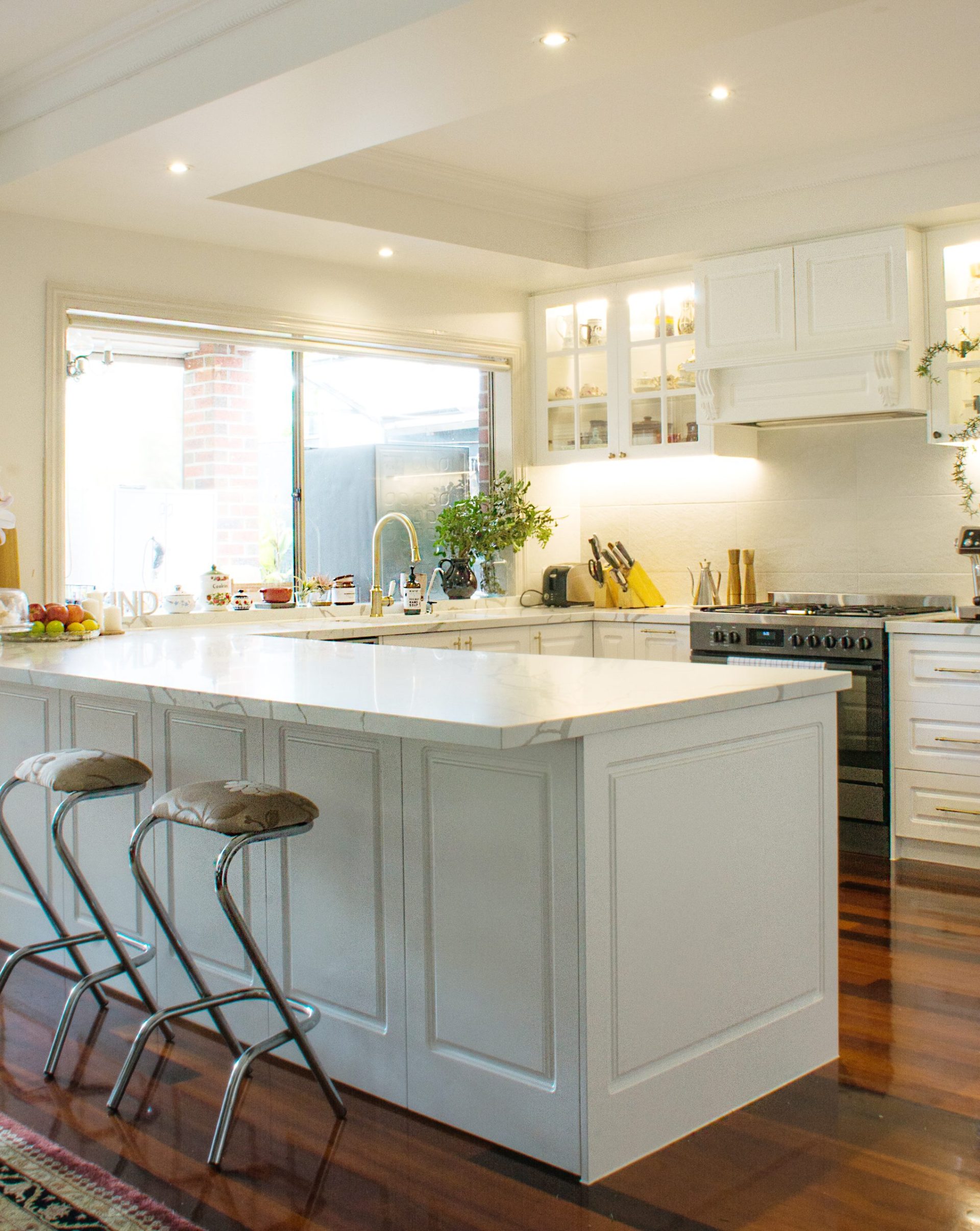 Bright kitchen with white cabinets, island, modern stove, and stools on wooden floor. Large window with natural light. Bright kitchen with white cabinets, island, modern stove, and stools on wooden floor. Large window with natural light.