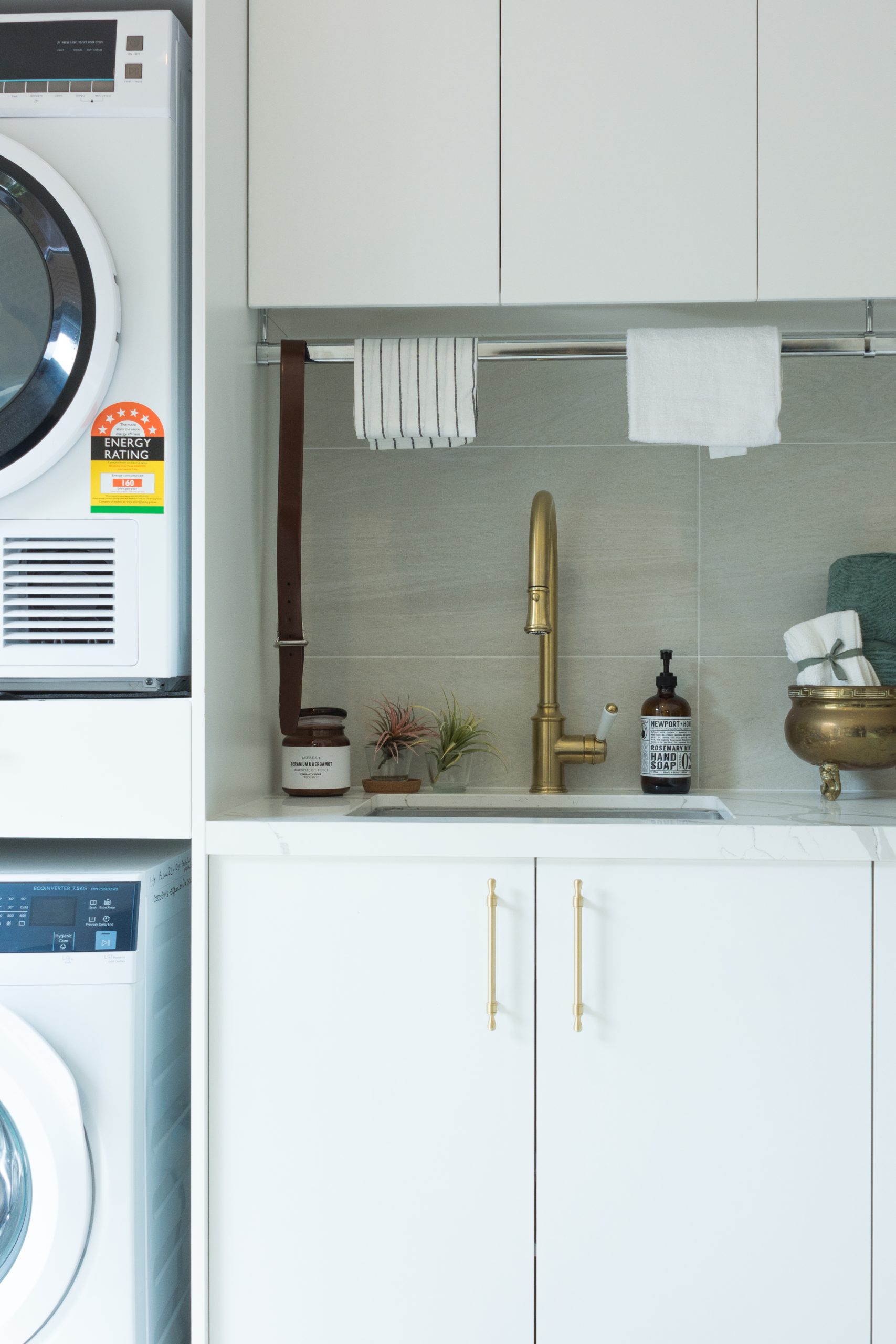 A laundry room with a stacked washer and dryer, gold faucet, and shelves holding towels and soap. A laundry room with a stacked washer and dryer, gold faucet, and shelves holding towels and soap.