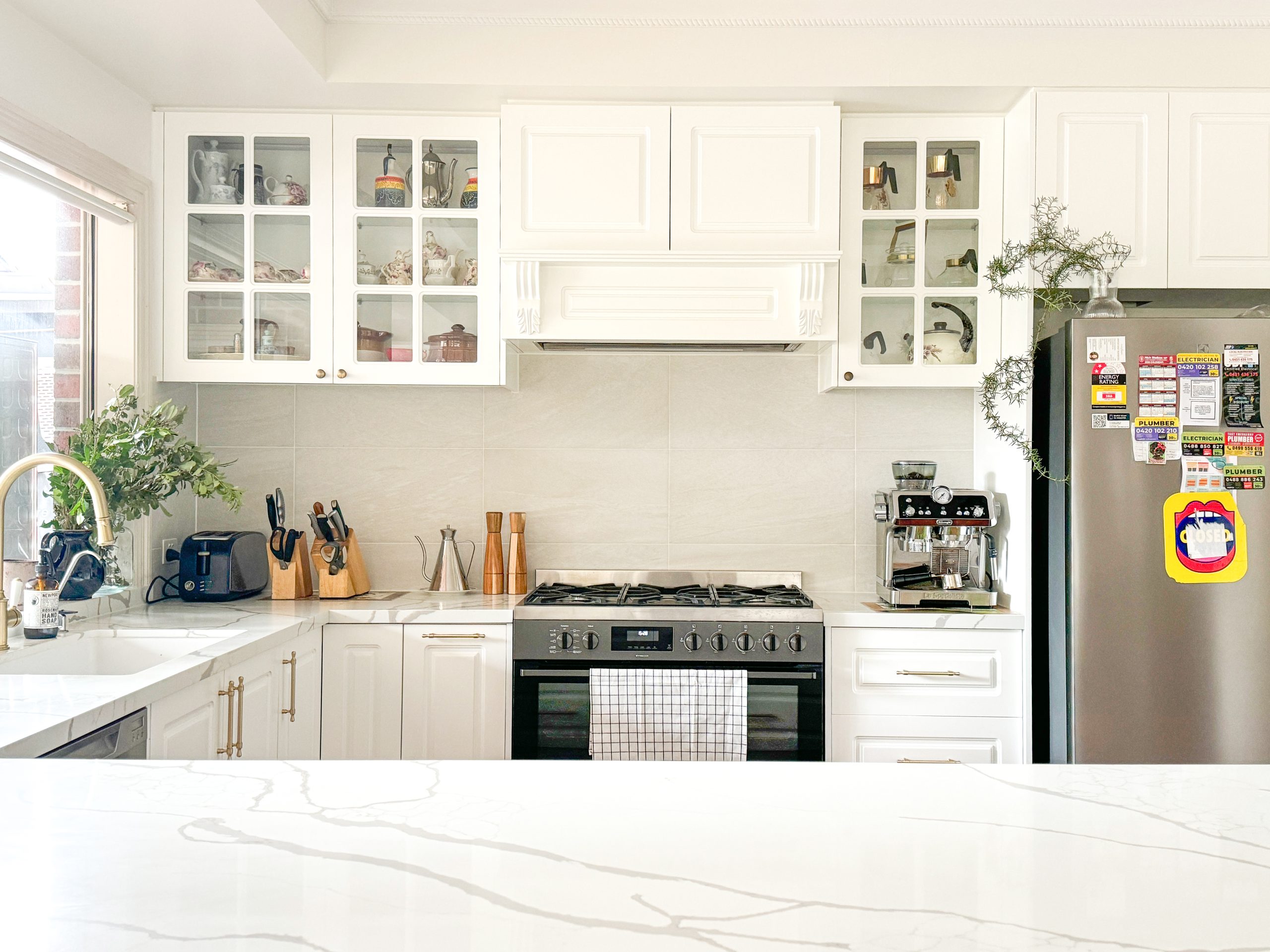 Bright kitchen with white cabinets, stainless steel appliances, and a marble countertop, decorated with plants.