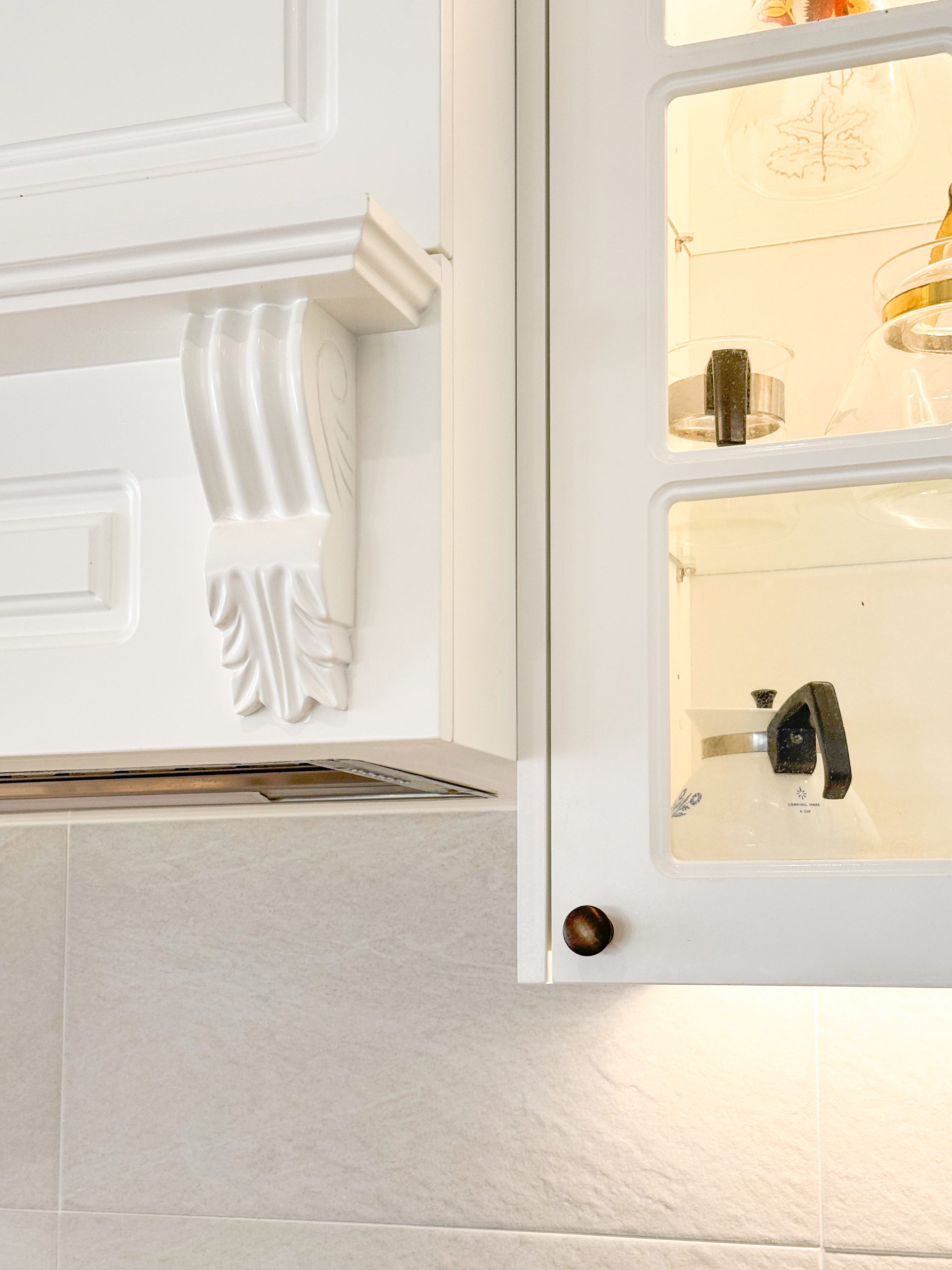 White kitchen cabinets with ornate details and glass doors, revealing cookware inside. Neutral tiled backsplash. White kitchen cabinets with ornate details and glass doors, revealing cookware inside. Neutral tiled backsplash.