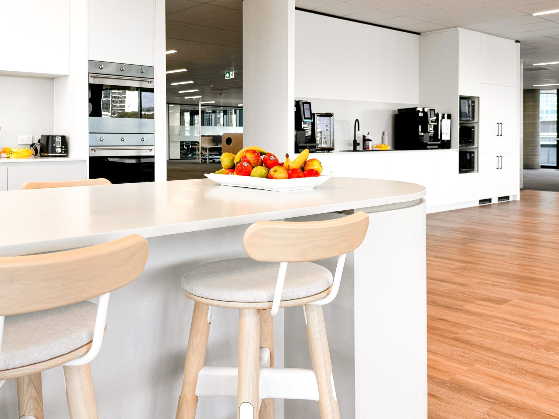 Modern kitchen with a curved island, wooden stools, fruit bowl, and sleek appliances on wooden flooring. Modern kitchen with a curved island, wooden stools, fruit bowl, and sleek appliances on wooden flooring.