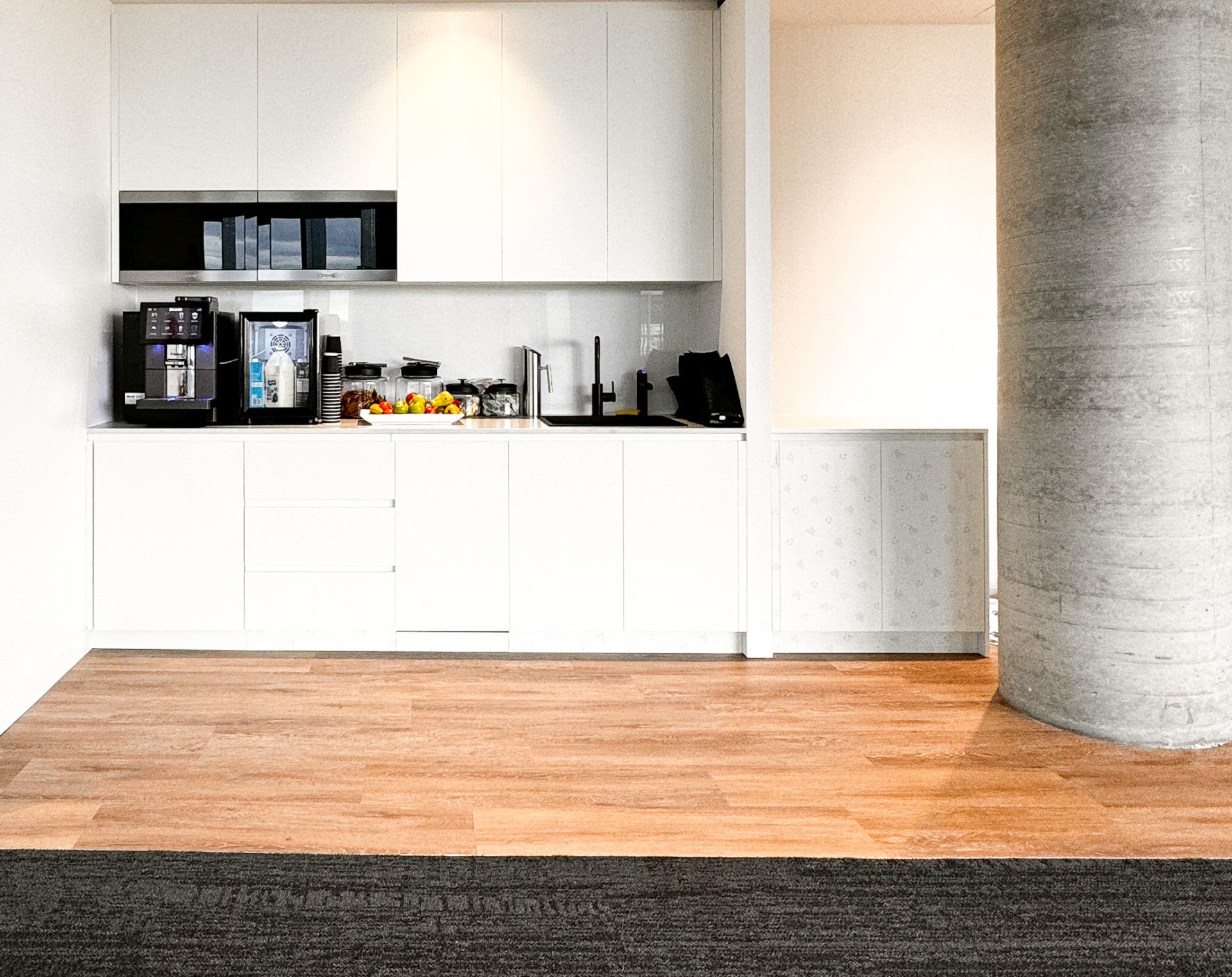 Modern kitchenette with white cabinets, coffee machine, fruit bowl, and a concrete pillar on a wood floor. Modern kitchenette with white cabinets, coffee machine, fruit bowl, and a concrete pillar on a wood floor.
