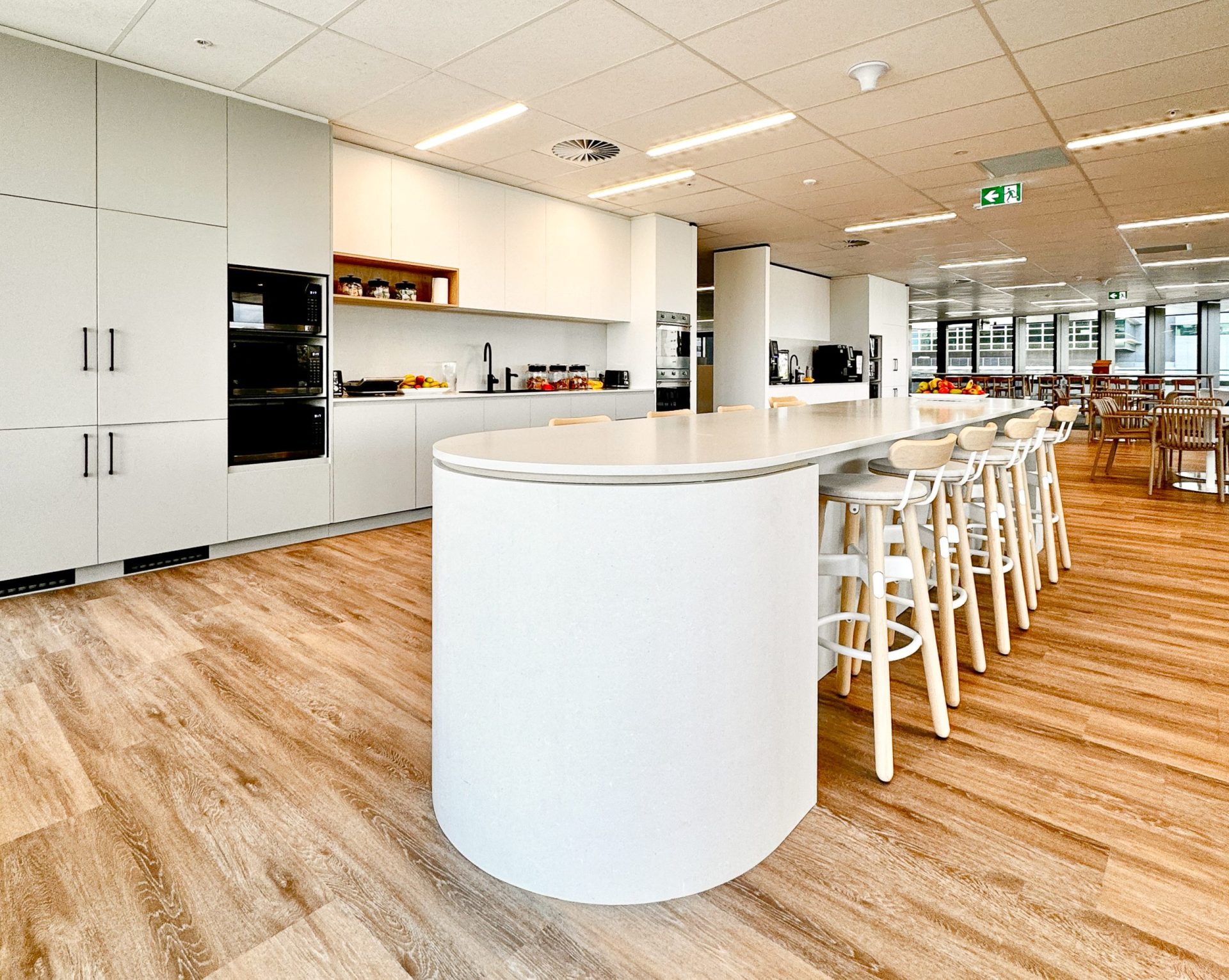 Modern office kitchen with white cabinets, a long island, bar stools, and wooden flooring. Bright with overhead lights. Modern office kitchen with white cabinets, a long island, bar stools, and wooden flooring. Bright with overhead lights.
