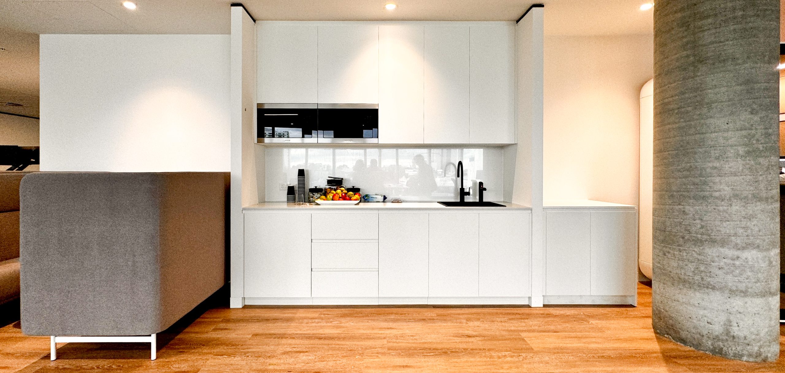 Modern white kitchenette with cabinets, a sink, a microwave, a fruit bowl, and a gray sofa on wooden floor.